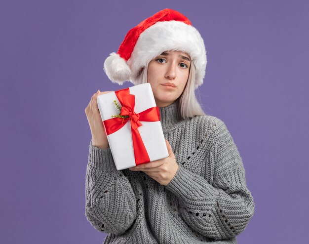 Junge blonde Frau in Winterpullover und Weihnachtsmütze, die ein Geschenk mit traurigem Gesichtsausdruck über lila Wand hält