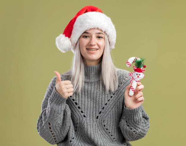 Junge blonde Frau in Winterpullover und Weihnachtsmütze mit Weihnachtszuckerstange lächelnd fröhlich mit Daumen nach oben stehend über grüner Wand