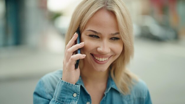 Junge blonde Frau lächelt selbstbewusst und spricht auf der Straße mit dem Smartphone