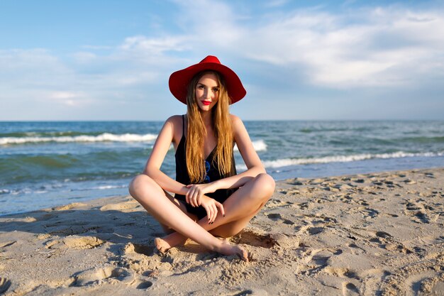 Junge blonde Frau meditieren in der Nähe von Ozean, Bohnenurlaub, Sonnenschein, tragen roten Hut und Balkenoberteil, gesunden Lebensstil, Yoga-Stimmung. Auf Sand sitzen und Urlaub genießen.