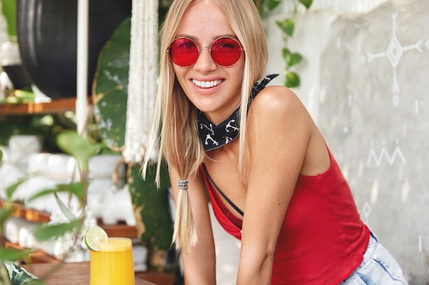 junge blonde Frau mit einer roten stilvollen Sonnenbrille, die in einem Café aufwirft