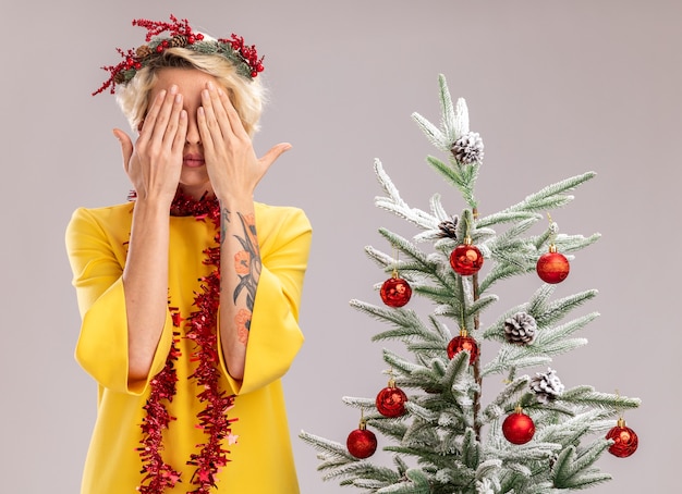 Junge blonde Frau mit Weihnachtskopfkranz und Lametta-Girlande um den Hals, die in der Nähe des geschmückten Weihnachtsbaums steht und die Augen mit den Händen isoliert auf weißer Wand bedeckt