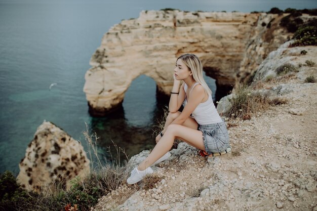 Junge blonde Frau sitzt auf einer Klippe und genießt den Blick auf das Meer