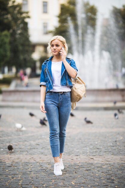 Junge blonde Mädchenfrau spricht Telefon auf Straßenwegquadrat-Fontain, gekleidet in Blue Jeans Suite mit Tasche auf ihrer Schulter in sonnigem Tag