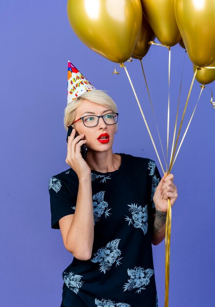 Junge blonde Partyfrau, die Brille und Geburtstagskappe hält, die Ballons hält, die Seite betrachten, die am Telefon lokalisiert auf lila Wand spricht