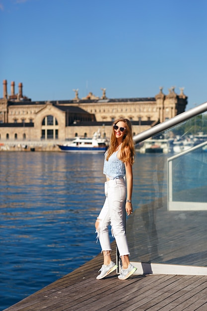 Junge blonde Reisen in Barcelona, stilvolle Kleidung und Sonnenbrille, atemberaubende Aussicht auf Meer und Architektur.