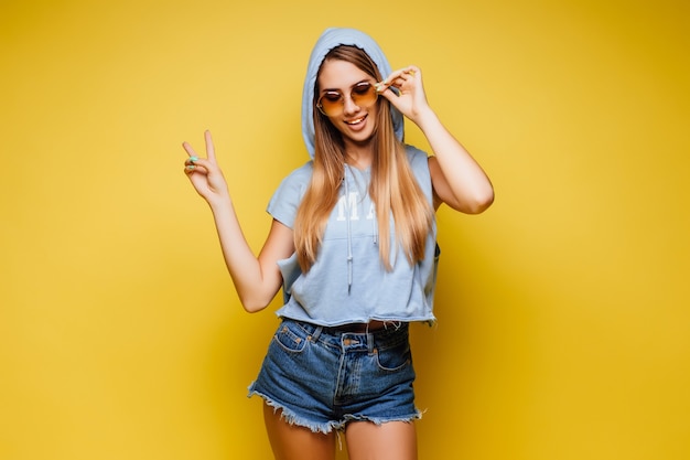 Junge blonde, süße Frau über isolierter gelber Wand in Sonnenbrille posiert