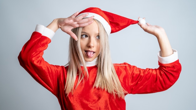 Junge blondhaarige Frau, die in einem Miss Weihnachtsmannkostüm auf dem grauen Wandhintergrund aufwirft