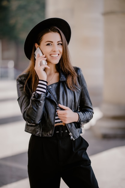 Junge braunhaarige Frau in einer Lederjacke, schwarzer Hut auf der Stadtpromenade haben ein Telefongespräch