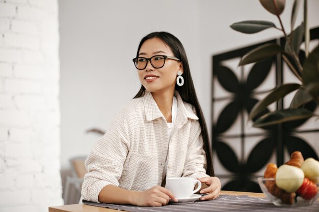 Junge brünette asiatische Frau in beiger stylischer Jacke posiert mit weißer Tasse Kaffee Attraktive Dame mit Brille lächelt, lehnt sich auf den Schreibtisch und hält Teeglas