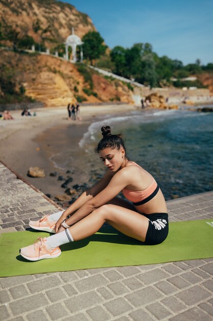Junge brünette Frau, die Fitness-Outfit trägt und auf Yogamatte draußen auf Pier im Sommer streckt.