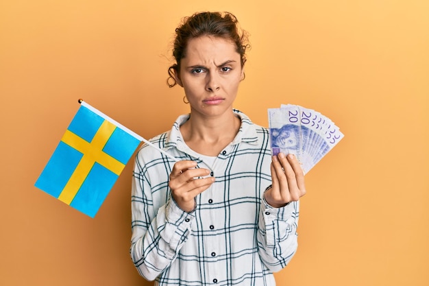 Kostenloses Foto junge brünette frau, die schwedenflagge und kronenbanknoten hält, skeptisch und nervös, stirnrunzelnd wegen problem. negativer mensch.