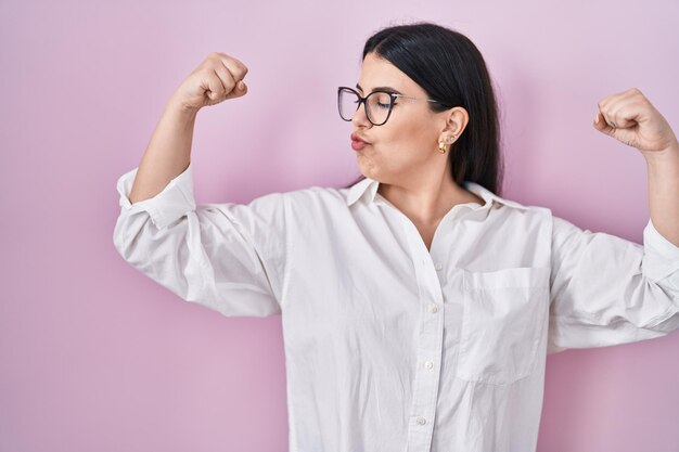 Junge brünette Frau, die über rosafarbenem Hintergrund steht und Armmuskeln zeigt, lächelt stolz Fitnesskonzept