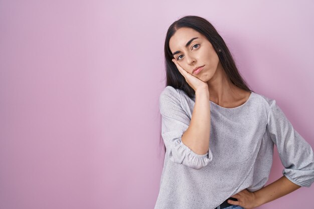 Junge brünette Frau, die über rosafarbenem Hintergrund steht und nachdenkt, sieht müde und gelangweilt von Depressionsproblemen mit verschränkten Armen aus.