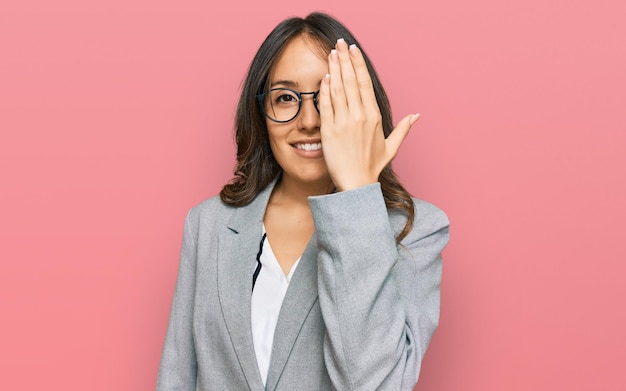 Junge brünette Frau in Business-Kleidung, die ein Auge bedeckt, mit selbstbewusstem Lächeln auf dem Gesicht und Überraschungsgefühl