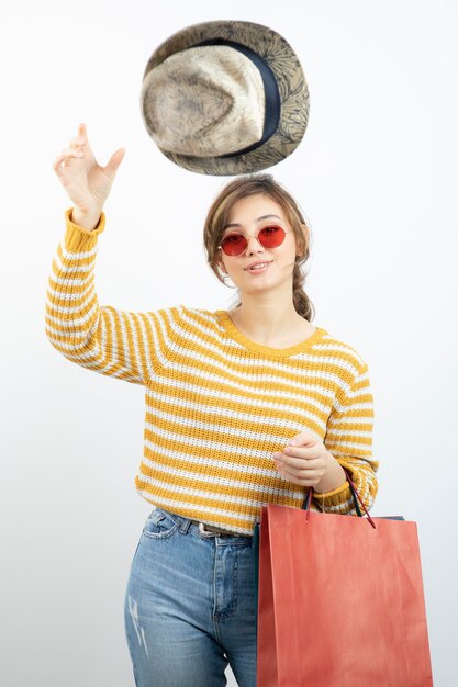 Junge brünette Frau mit Sonnenbrille, die Einkaufstüten hält und Hut hochwirft. Foto in hoher Qualität