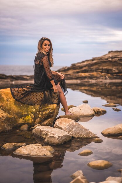 Junge brünette kaukasische Frau in einem langen schwarzen transparenten Kleid auf einigen Felsen in der Nähe des Meeres