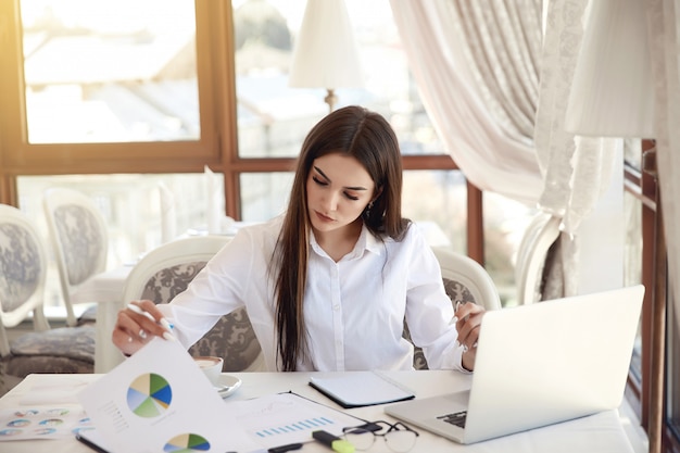 Junge Brunettegeschäftsdame analysiert Diagramme und arbeitet an dem Laptop