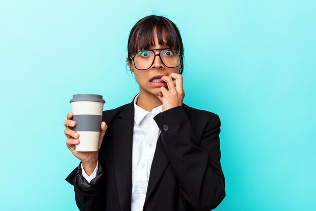 Junge business-mixed-race-frau, die einen kaffee hält, isoliert auf blauem hintergrund, fingernägel beißend, nervös und sehr ängstlich.