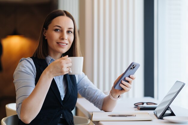 Junge charmante Frau, die mit Smartphone eine SMS schreibt, während sie allein im Kaffeegeschäft sitzt, das Kaffee trinkt, Gespräch mit Handy hat
