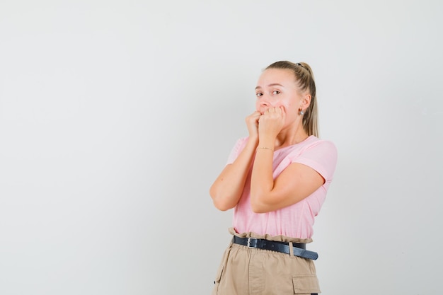 Junge Dame beißt emotional Fäuste in T-Shirt, Hose und sieht verängstigt aus. Vorderansicht.