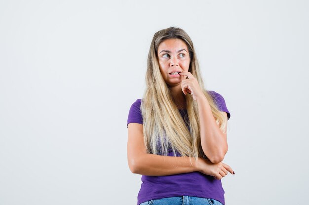 Junge Dame beißt Finger im violetten T-Shirt, Jeans und schaut ängstlich, Vorderansicht.