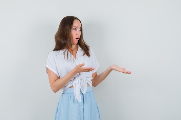 Junge Dame breitete ihre offenen Handflächen zur Seite, um etwas in weißer Bluse, blauem Rock zu zeigen und verblüfft auszusehen