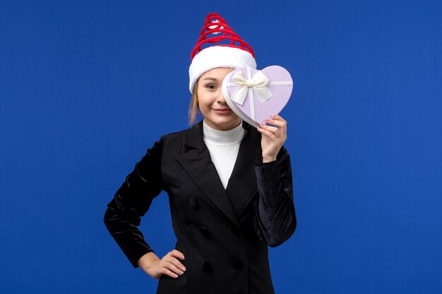 Junge Dame der Vorderansicht, die herzförmiges Geschenk auf Neujahrs-Weihnachtsgeschenk der blauen Wand hält