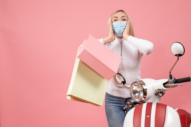 Junge Dame der Vorderansicht mit Maske, die Einkaufstaschen hält, die nahe Moped stehen