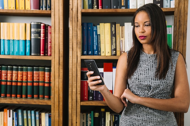 Junge Dame des Afroamerikaners, der Smartphone nahe Büchern verwendet