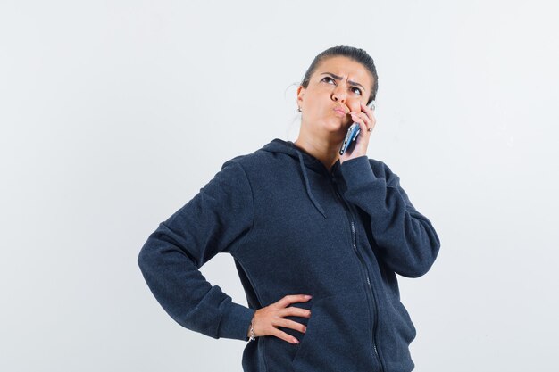 Junge Dame, die am Telefon spricht, während sie in Jacke denkt und verwirrt schaut. Vorderansicht.