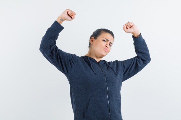 Junge Dame, die Arme hebt, die Siegergeste in der Jacke zeigt und flexibel schaut. Vorderansicht.