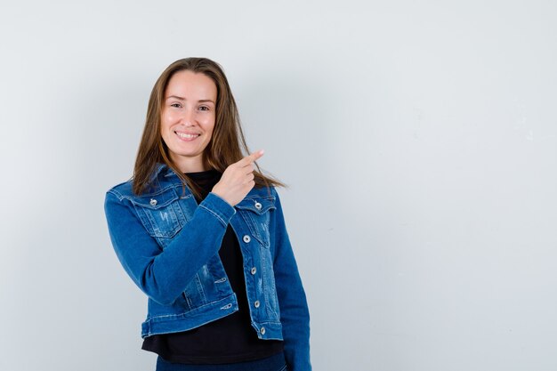 Junge Dame, die auf die obere rechte Ecke in Bluse, Jacke zeigt und fröhlich aussieht. Vorderansicht.