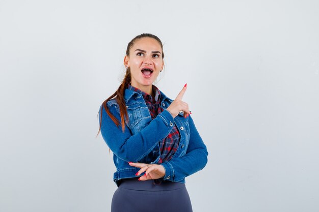 Junge Dame, die auf die obere rechte Ecke in kariertem Hemd, Jeansjacke zeigt und erstaunt aussieht, Vorderansicht.