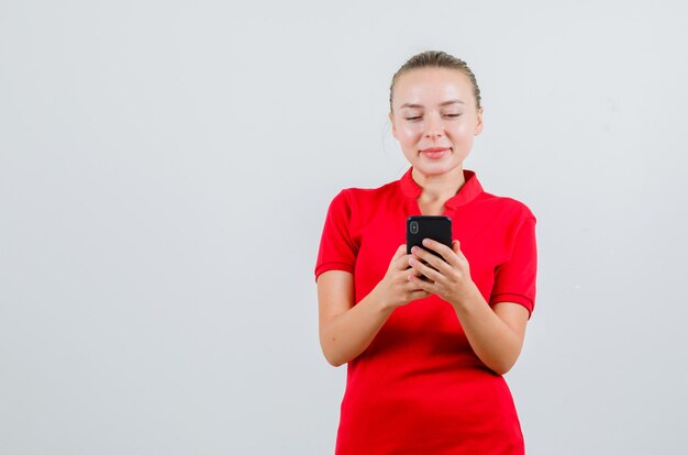 Junge Dame, die auf Handy in rotem T-Shirt tippt und fröhlich schaut