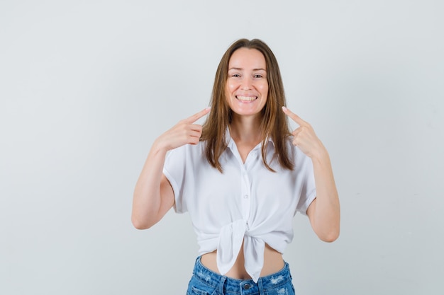 Junge Dame, die auf ihr Lächeln in der weißen Bluse zeigt und lustig schaut. Vorderansicht.