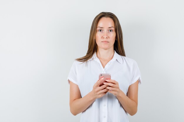 Junge Dame, die beim Halten des Telefons in der weißen Bluse steht und ernst schaut