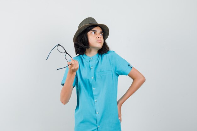 Junge Dame, die beiseite schaut, während sie ihre Brille im blauen Hemd hält und verwirrt schaut