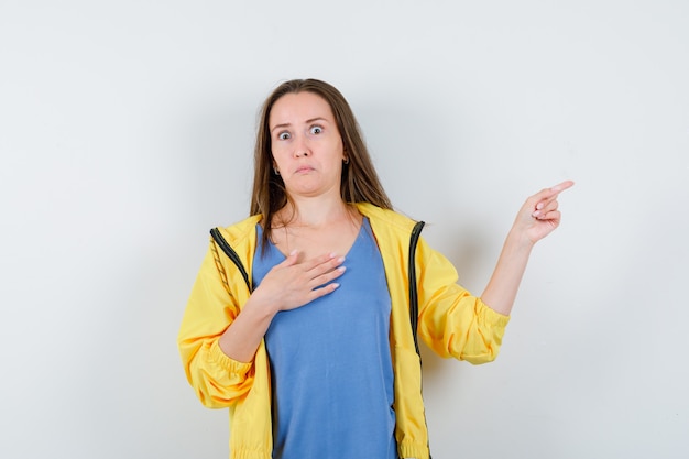 Junge Dame, die beiseite zeigt, die Hand auf der Brust in T-Shirt, Jacke hält und verängstigt aussieht. Vorderansicht.