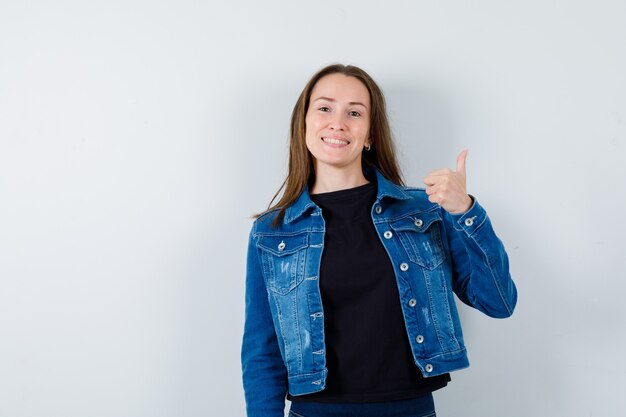 Junge Dame, die Daumen in Bluse, Jacke zeigt und fröhlich aussieht. Vorderansicht.