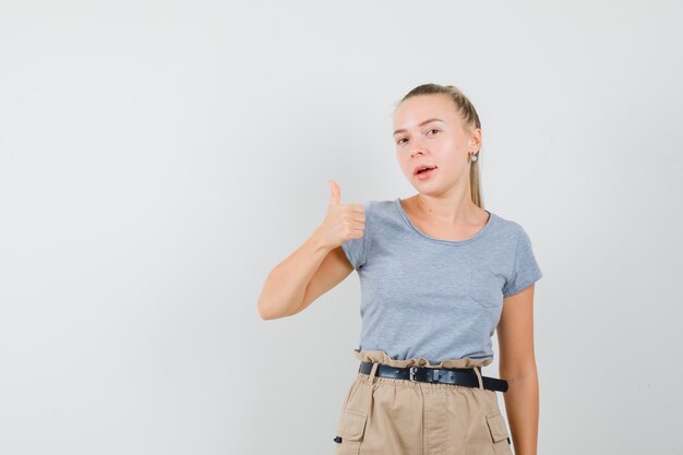 Junge Dame, die Daumen in T-Shirt und in der Hose zeigt und erfreut aussieht