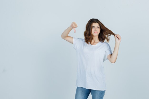 Junge Dame, die Daumen nach unten zeigt, während Haarsträhne im T-Shirt, in den Jeans hält und unzufrieden aussieht, Vorderansicht.