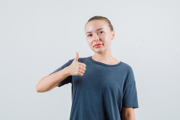 Junge Dame, die Daumen oben im grauen T-Shirt zeigt und erfreut schaut