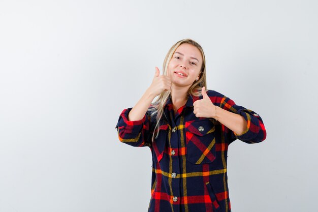 Junge Dame, die Daumen oben im karierten Hemd zeigt und glückliche Vorderansicht schaut.