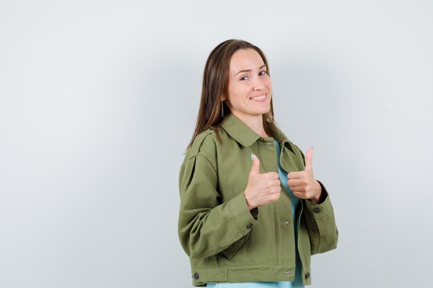 Junge Dame, die doppelte Daumen in grüner Jacke zeigt und fröhlich aussieht, Vorderansicht.