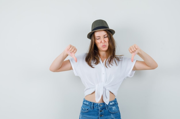 Junge Dame, die doppelte Daumen nach unten in T-Shirt, Jeans, Hut zeigt und unzufrieden aussieht, Vorderansicht.