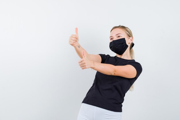 Junge Dame, die doppelte Daumen oben im schwarzen T-Shirt, in der Maske zeigt und glücklich schaut. Vorderansicht.