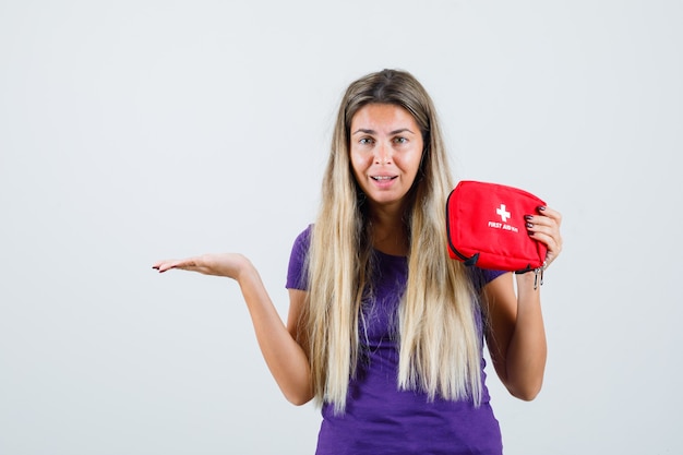 Junge Dame, die Erste-Hilfe-Kit hält, Handfläche beiseite in violettem T-Shirt, Vorderansicht spreizend.