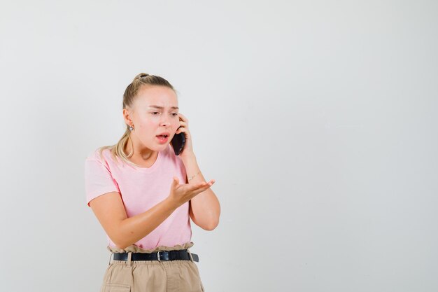 Junge Dame, die etwas auf Handy in T-Shirt, Hosen bespricht und verwirrt, Vorderansicht schaut.