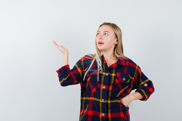 Junge Dame, die etwas zeigt, während sie Hand in der Taille im karierten Hemd hält und selbstbewusst aussieht. Vorderansicht.